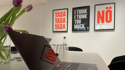 Ein Laptop steht auf einem Tisch. Im Hintergrund sind drei Poster an der Wand.